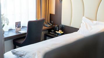 Ein Hotelzimmer mit einem Bett, einem Schreibtisch mit Telefon, einem Sessel, einem kleinen Tisch mit Lampe und einem Fenster mit Vorhängen.