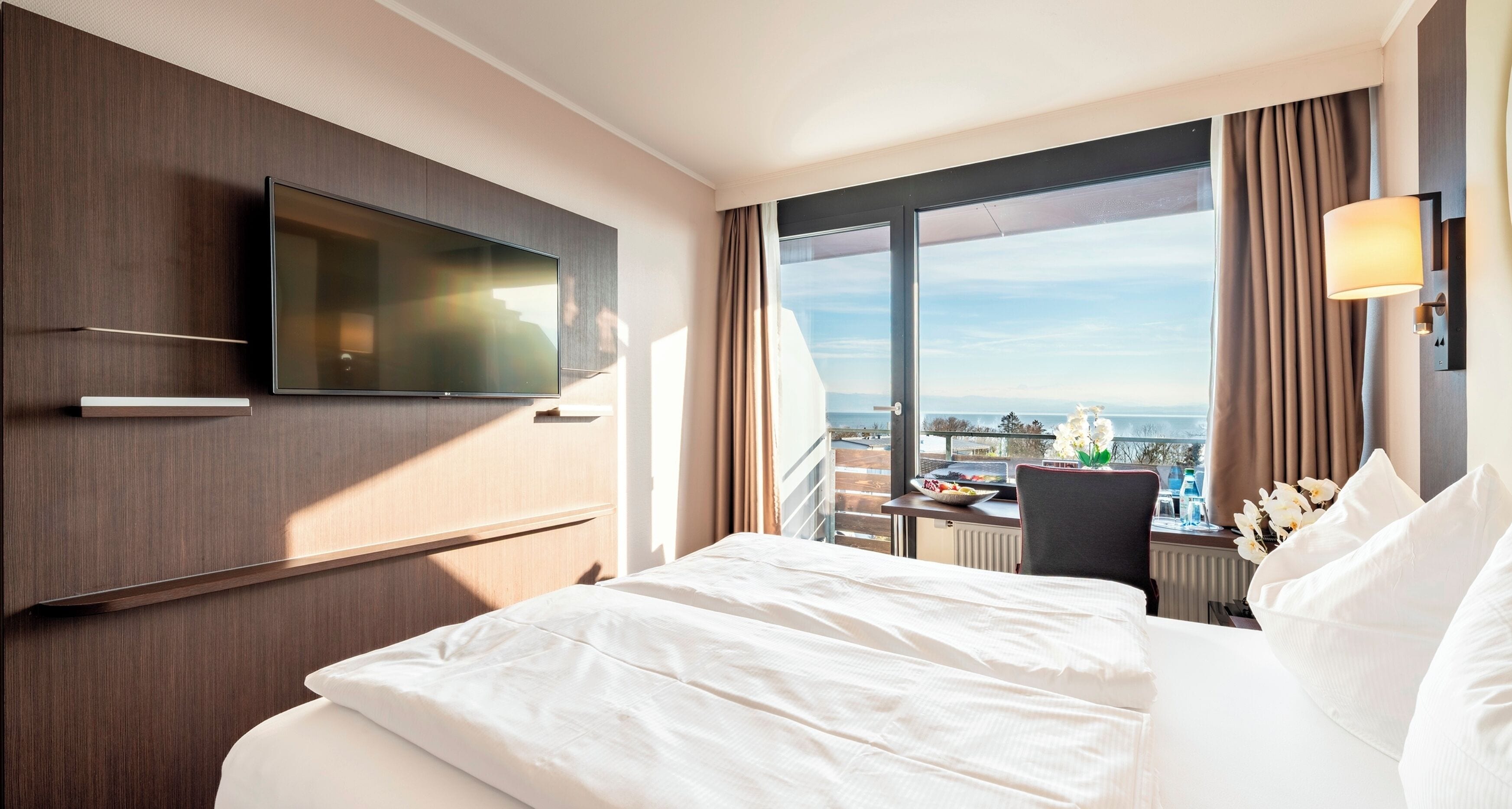 Une chambre d’hôtel dotée d’un grand lit, d’un téléviseur à écran plat fixé au mur, d’un bureau avec une chaise et offrant une vue sur la ville grâce à de grandes fenêtres.