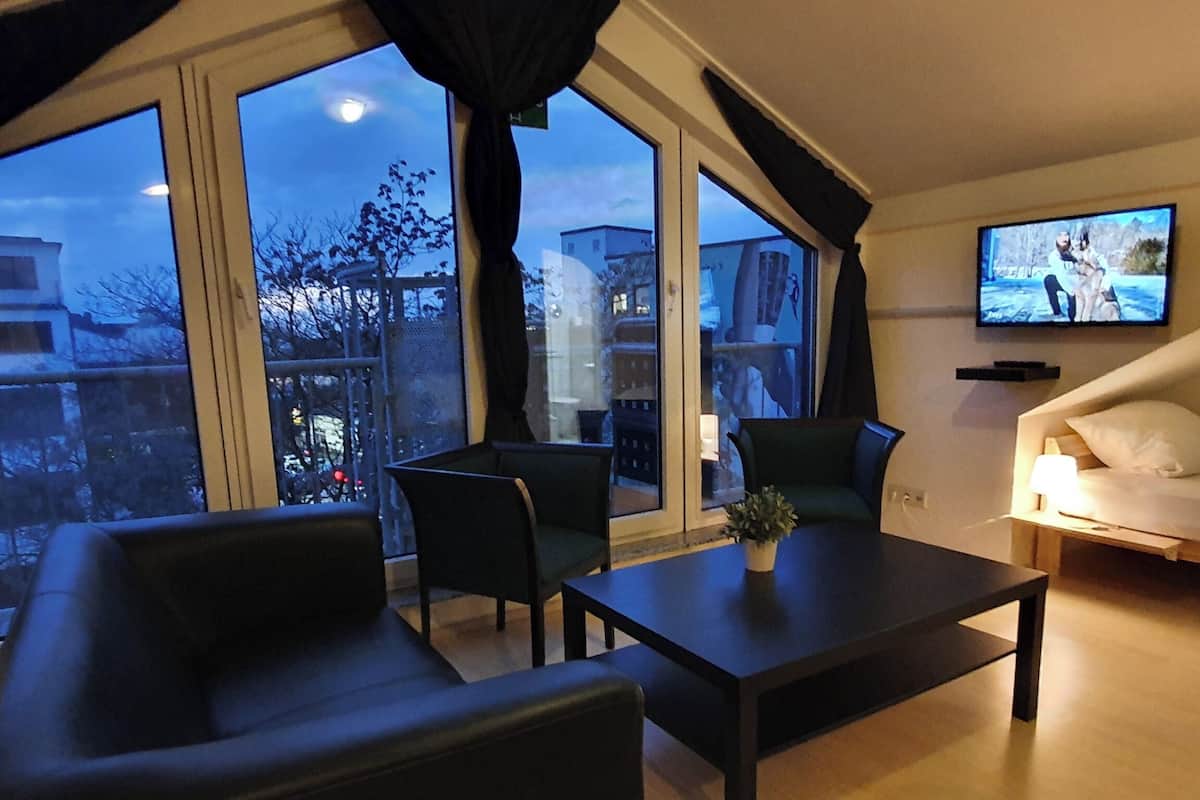 A modern living room with a large window, a flat-screen TV mounted on the wall, and a cozy seating area with a sofa and chairs.