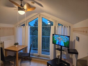 A room with a television, a ceiling fan, a table with a chair, and a window with curtains.