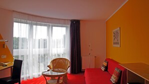A room with a red sofa, a wicker chair, a small table, a desk with a chair, a window with sheer curtains, and a radiator. - Hotel am Buschkrugpark (Berlin)
