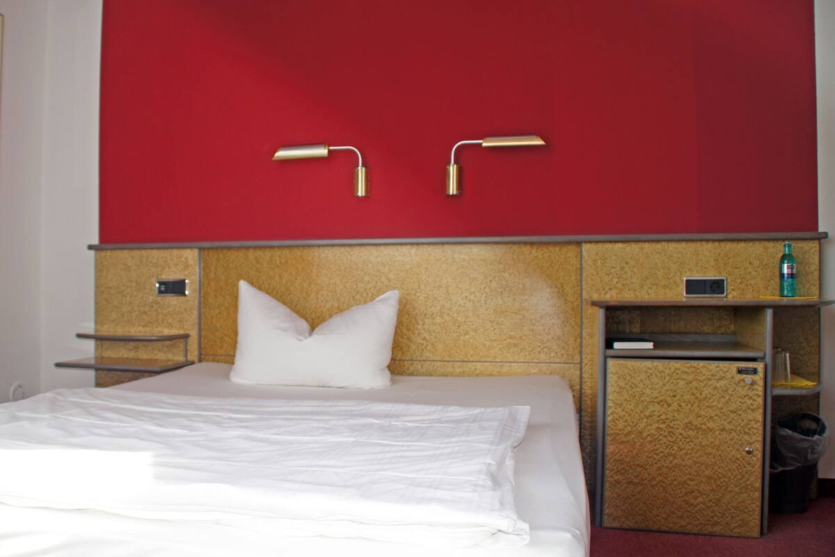 A hotel room with a bed, a red wall, a nightstand, and a small shelf with a bottle.