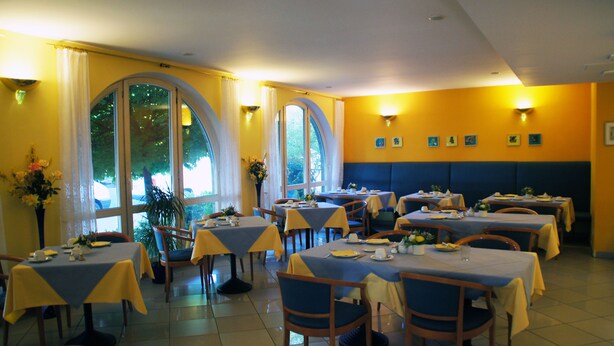 A restaurant with multiple tables and chairs, large windows, and a yellow and blue color scheme. - Hotel am Buschkrugpark (Berlin)