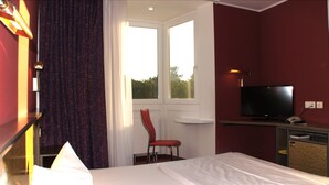 A hotel room with a bed, a red chair, a television, and a window with curtains. - Hotel am Buschkrugpark (Berlin)