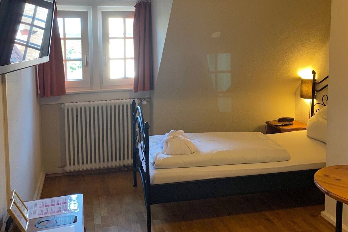 A small, cozy room with a bed, a desk with a computer, a window with curtains, and a radiator.
