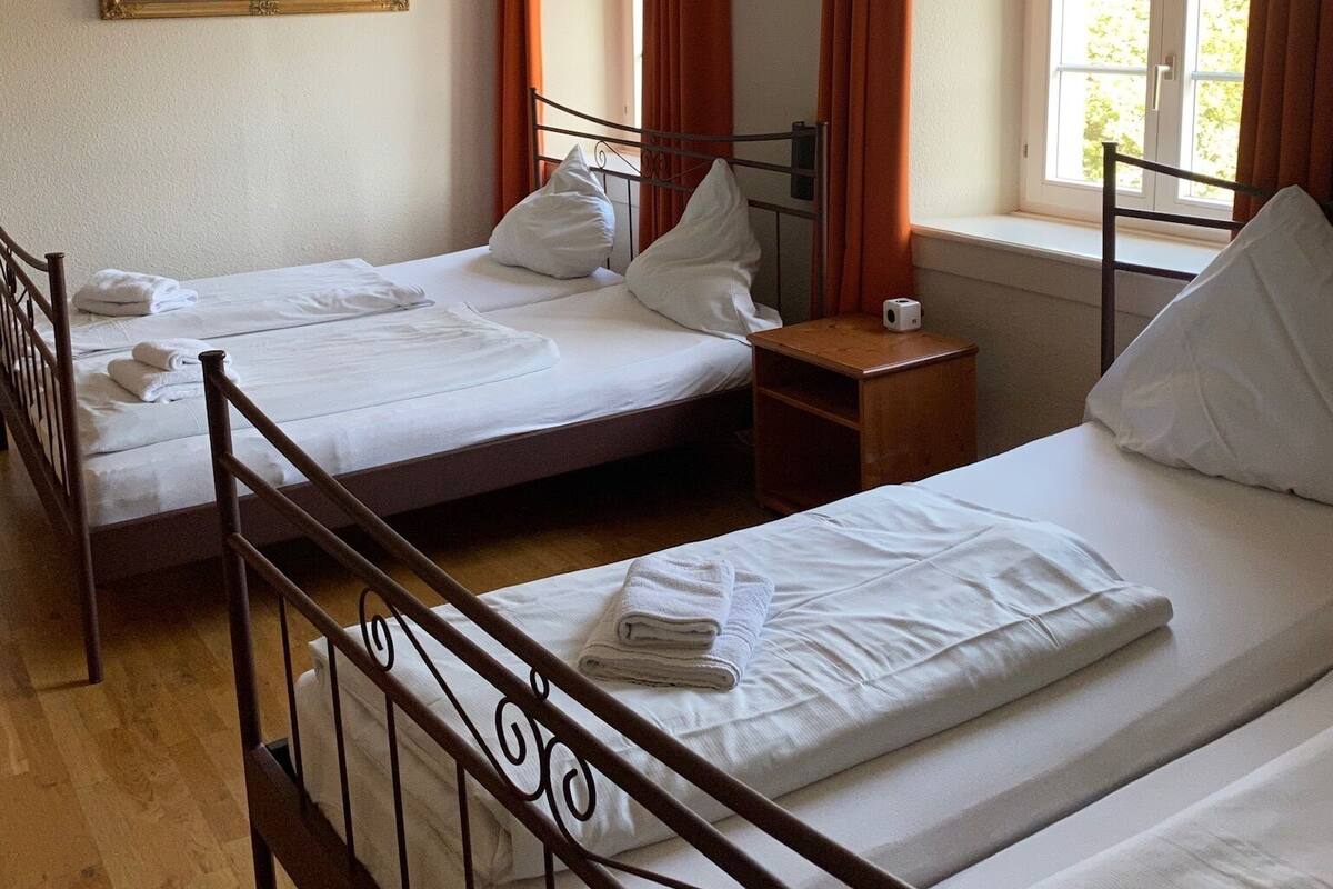 A hotel room with two beds, a wooden floor, a mirror, and a window with curtains.