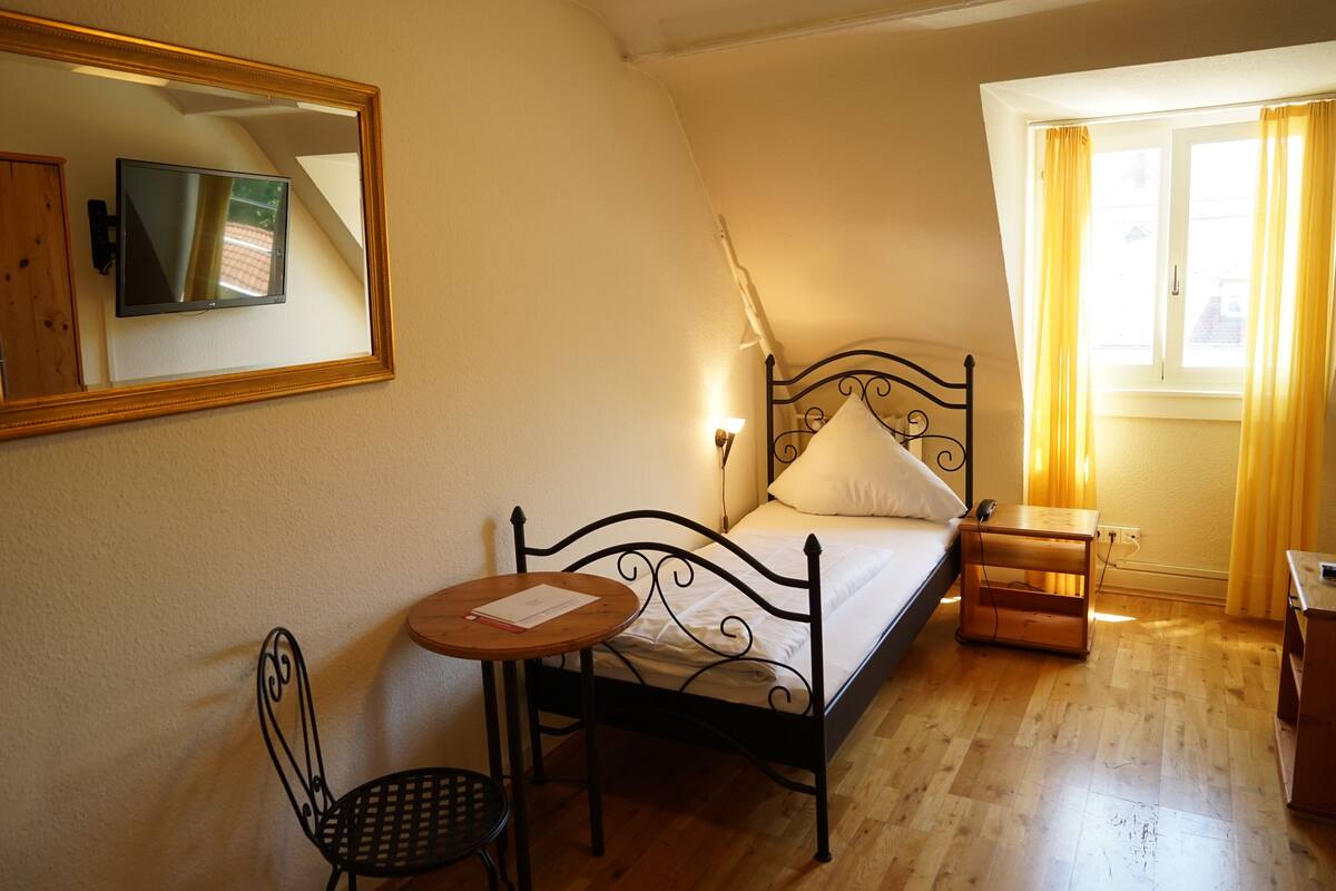 A bedroom with a bed, a bedside table, a chair, a small table, a mirror, and a window with curtains.