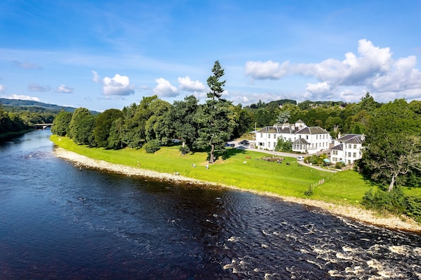 A large, white, multi-story building with a prominent roof, situated beside a river. - Banchory Lodge Hotel (Banchory)