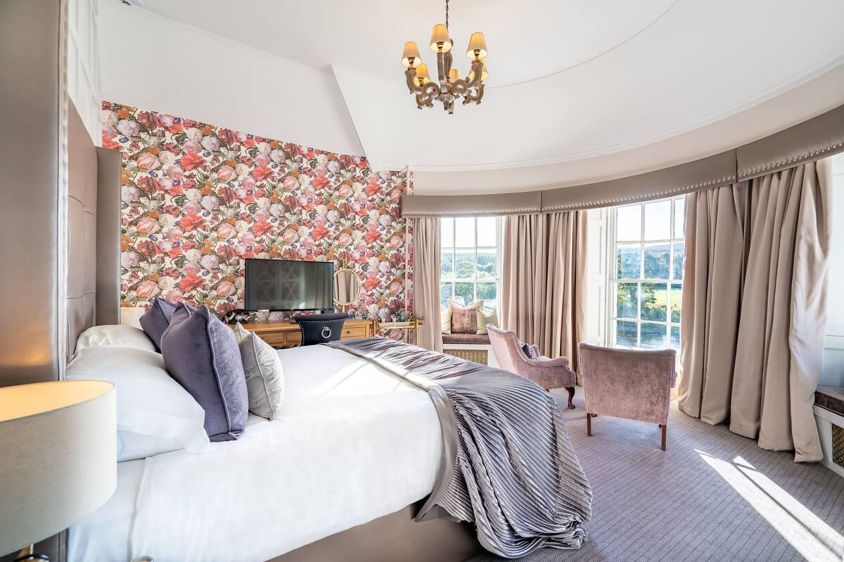 A spacious bedroom with a large bed, a television, a desk, and a chair. The room has a floral wallpaper and a view of the outdoors.