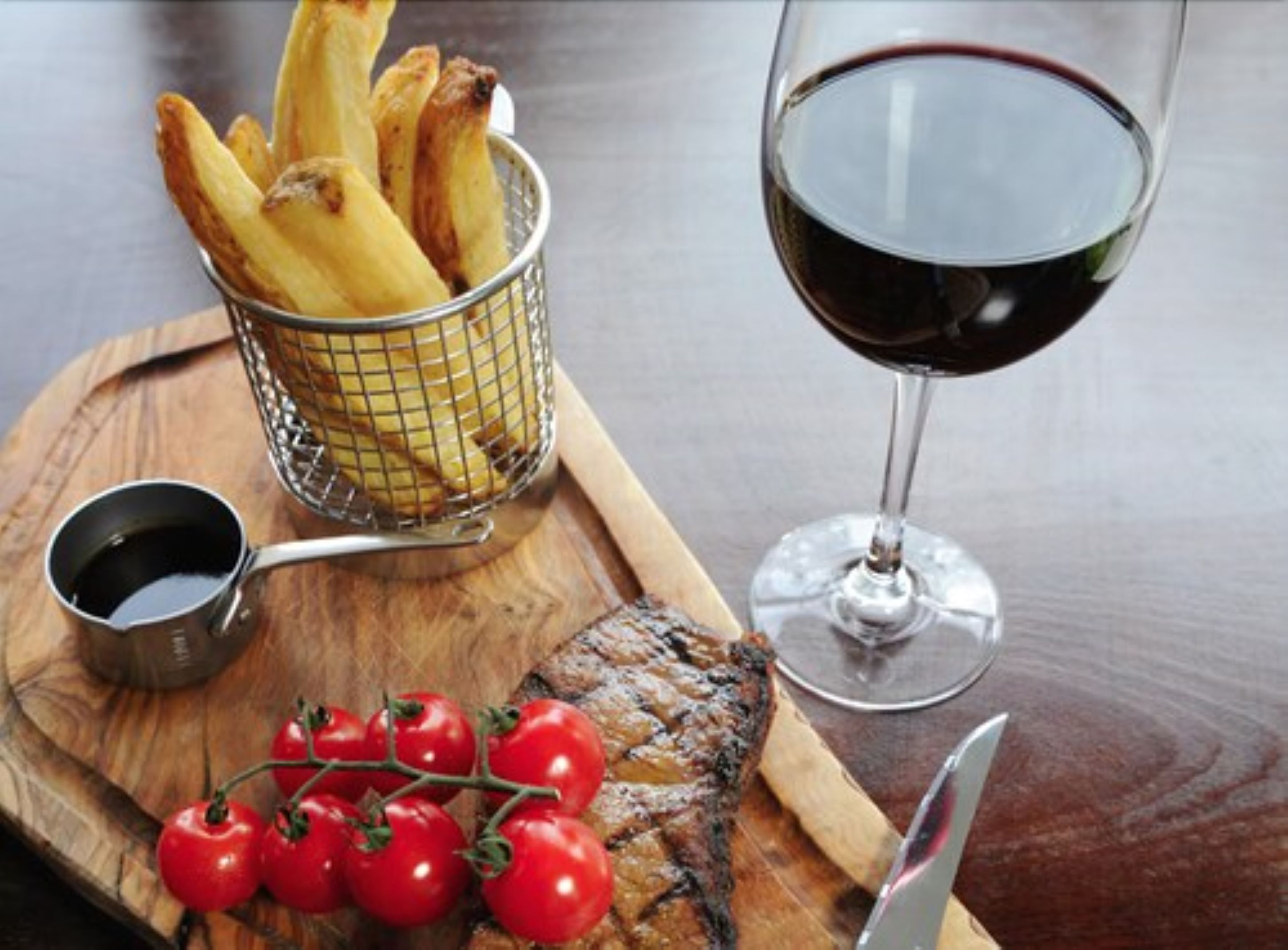 A wooden cutting board with a basket of fries, a glass of red wine, a small container of sauce, and a knife.