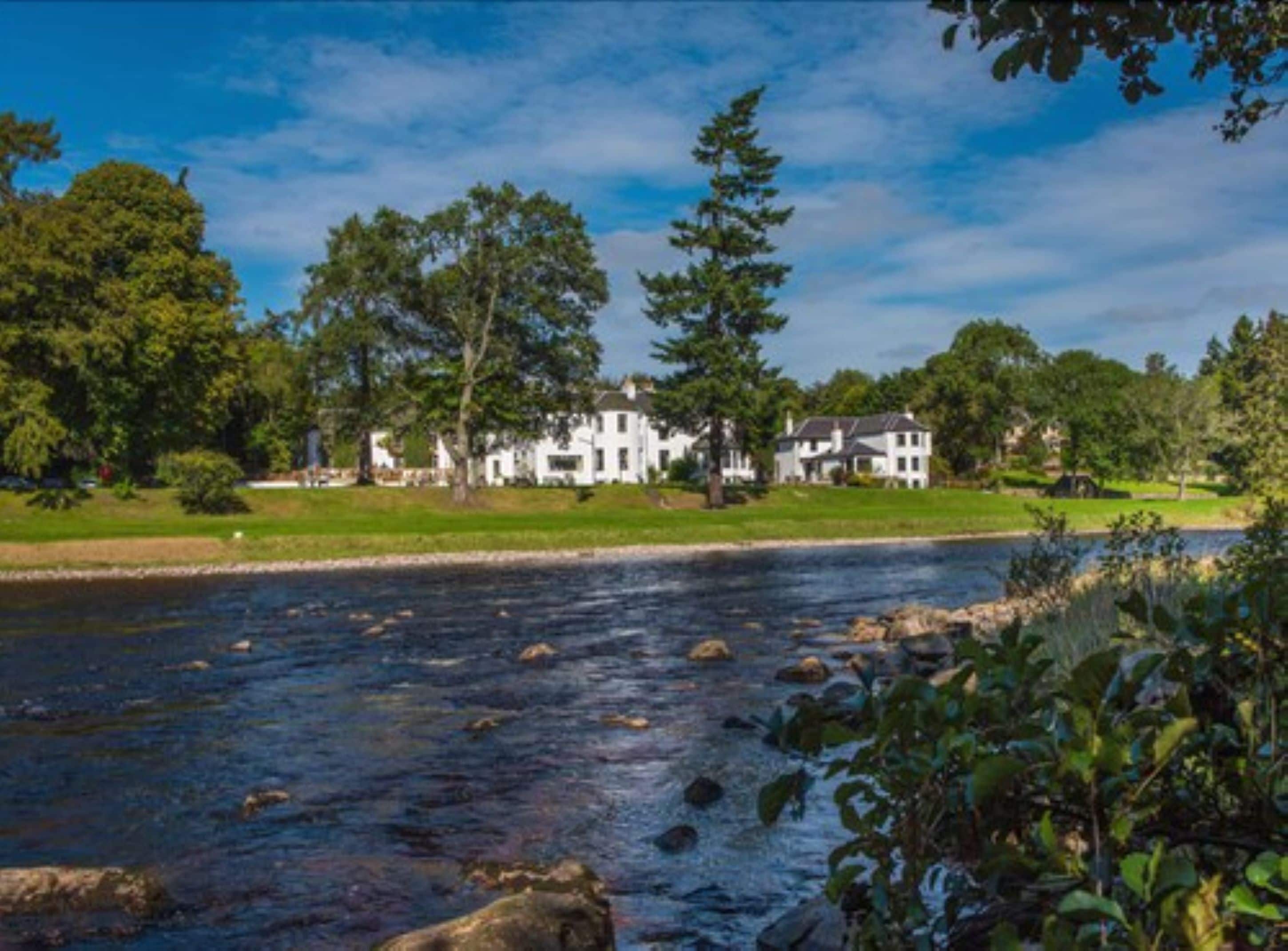 A riverside view of a large white house with multiple buildings, surrounded by lush greenery and trees.