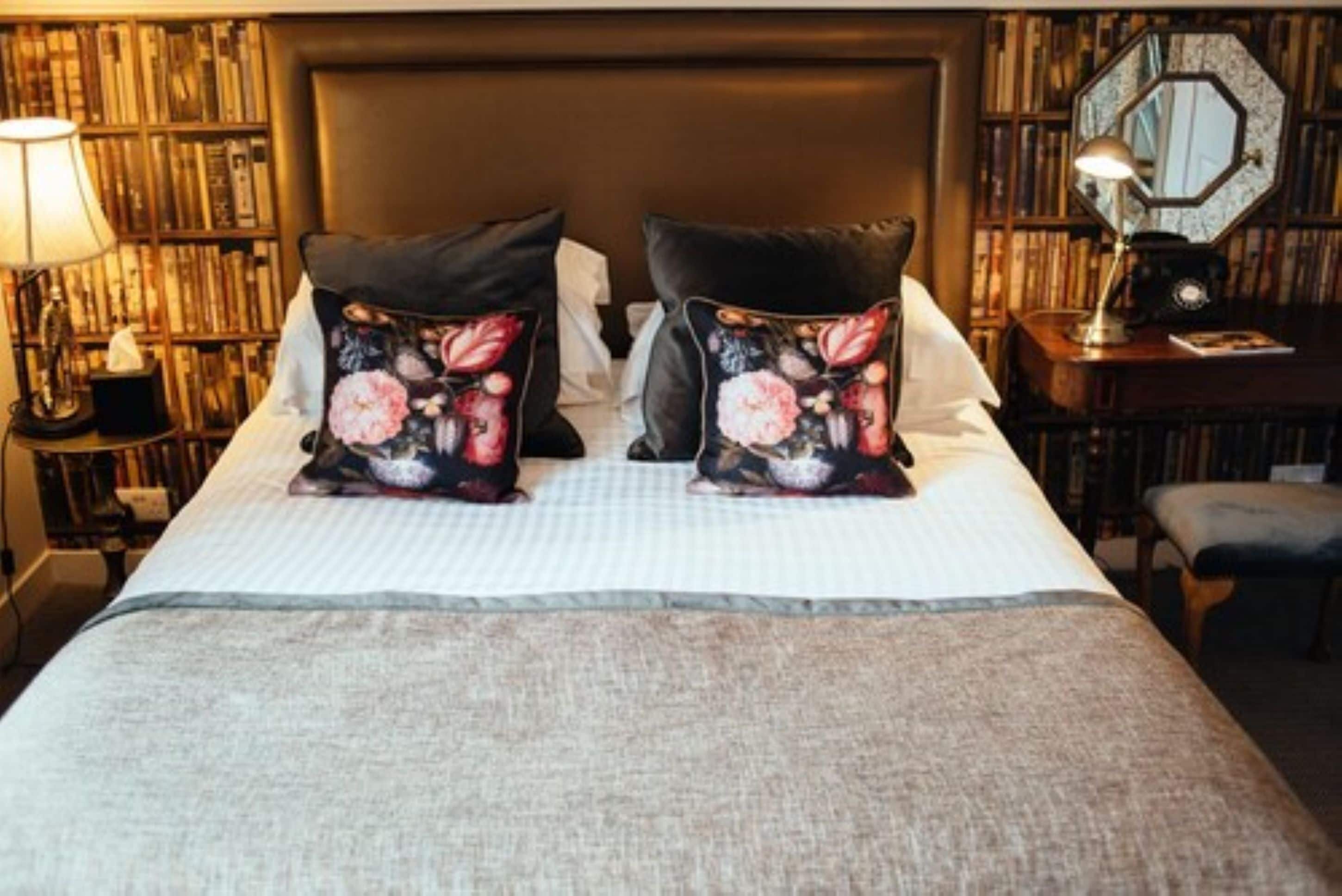A neatly made bed with floral pillows, a bedside table with a lamp, and a bookshelf in the background.