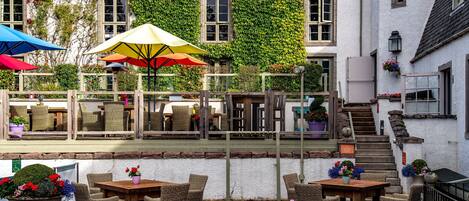 An outdoor seating area with wicker furniture and umbrellas, a staircase leading to a building with ivy-covered walls, and flower planters.