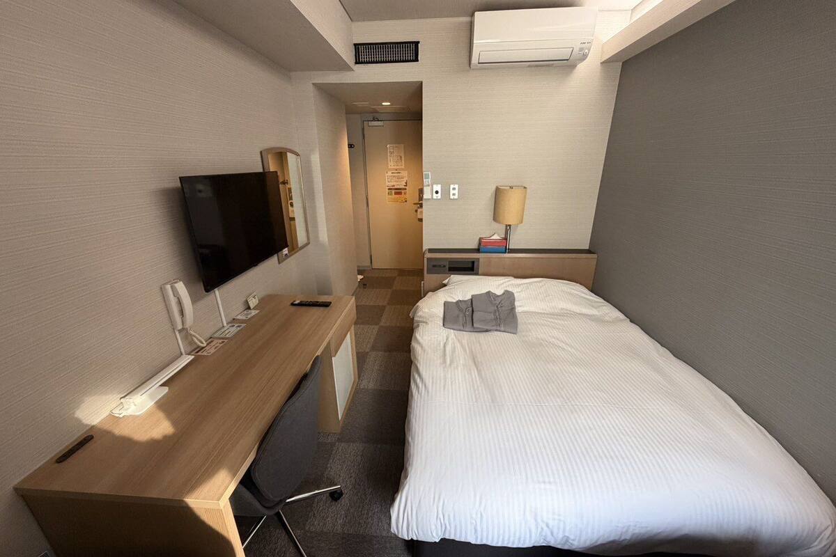 Habitación de hotel con cama, escritorio con silla, televisor, aire acondicionado y lámpara.