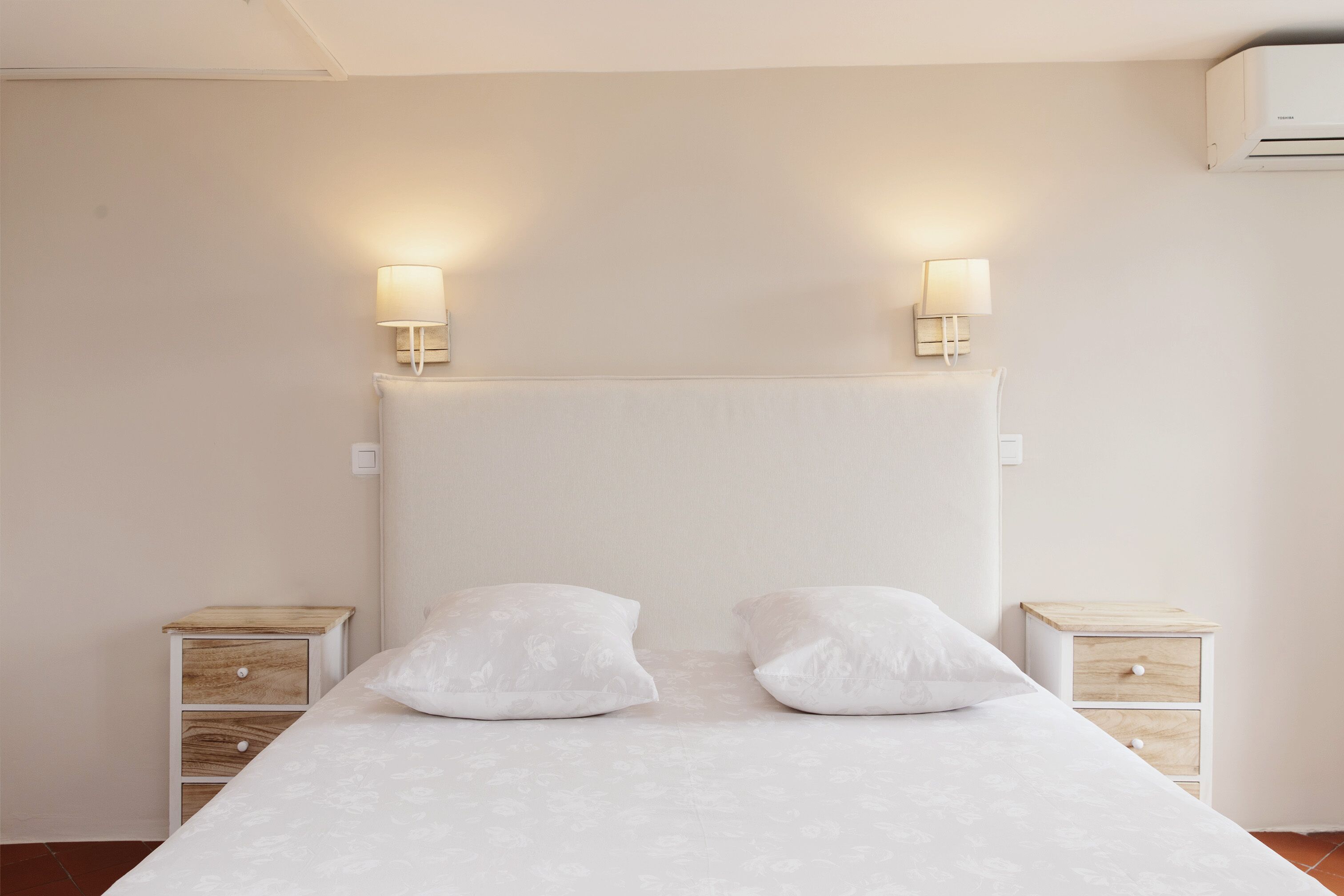 Un lit bien fait, avec deux oreillers, deux tables de chevet dotées de tiroirs et des lampes fixées au mur.