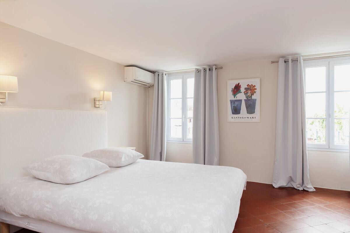 A bedroom with a large bed, two wall-mounted lamps, a window with curtains, and a painting on the wall.