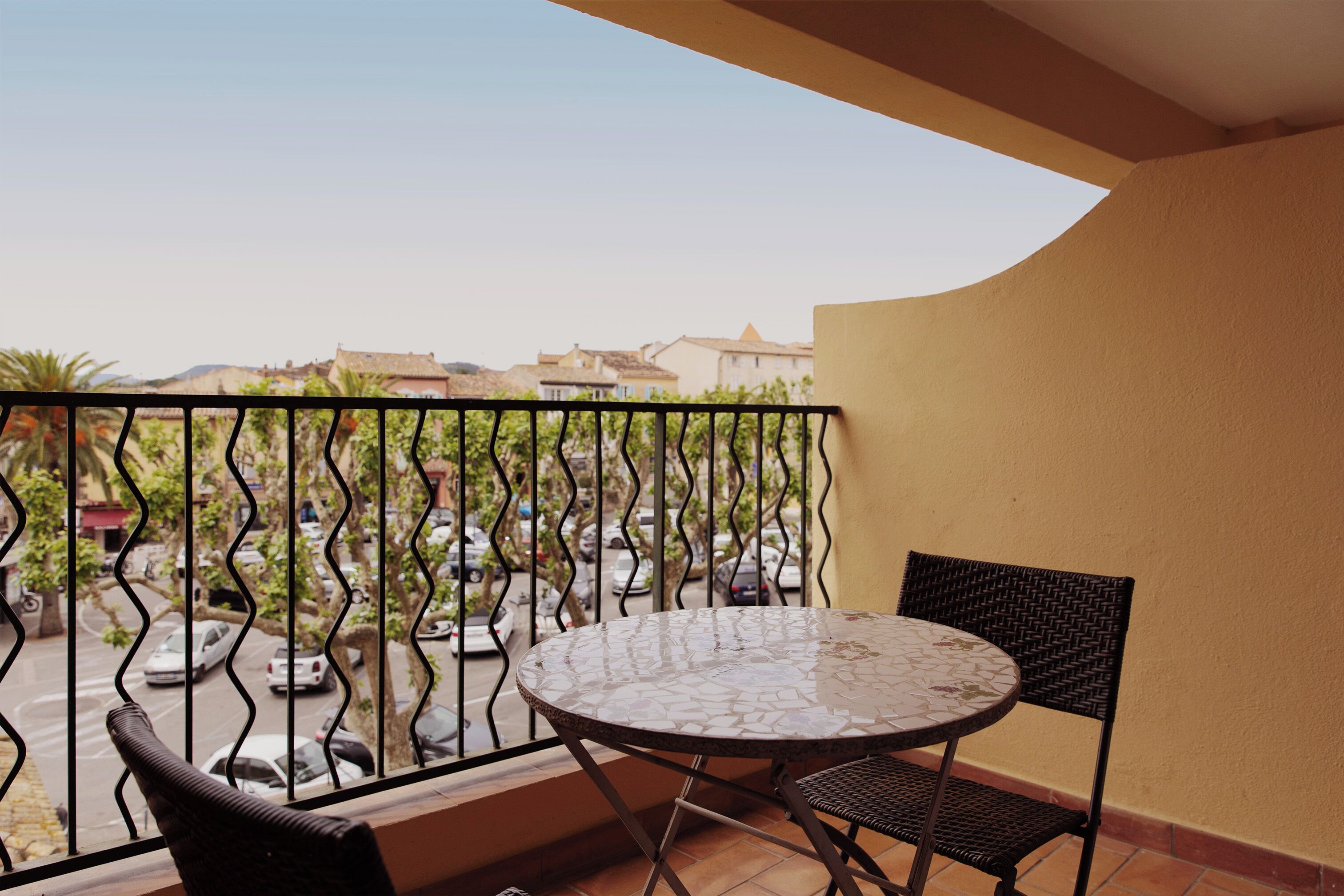 A balcony with a round table and two chairs, overlooking a street with parked cars and buildings.