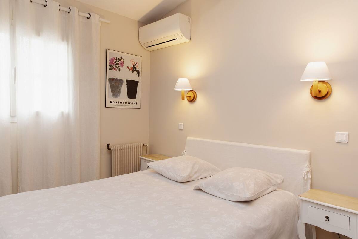 A bedroom with a bed, two bedside tables, an air conditioning unit, a radiator, and a wall-mounted lamp.