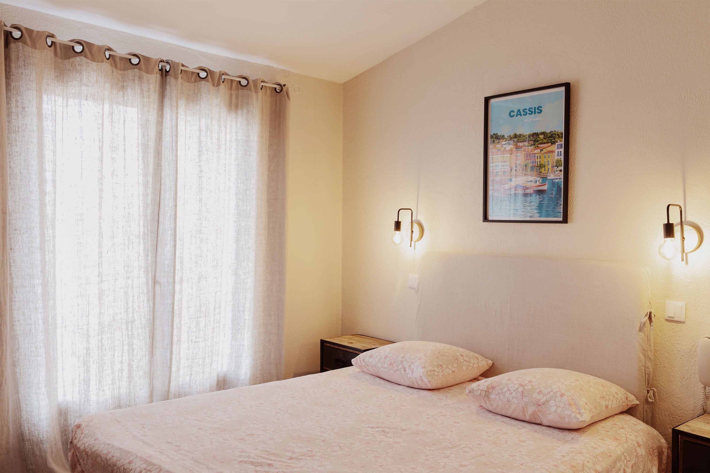 A bedroom with a bed, two nightstands, a wall-mounted light, and a framed picture of Cassis.