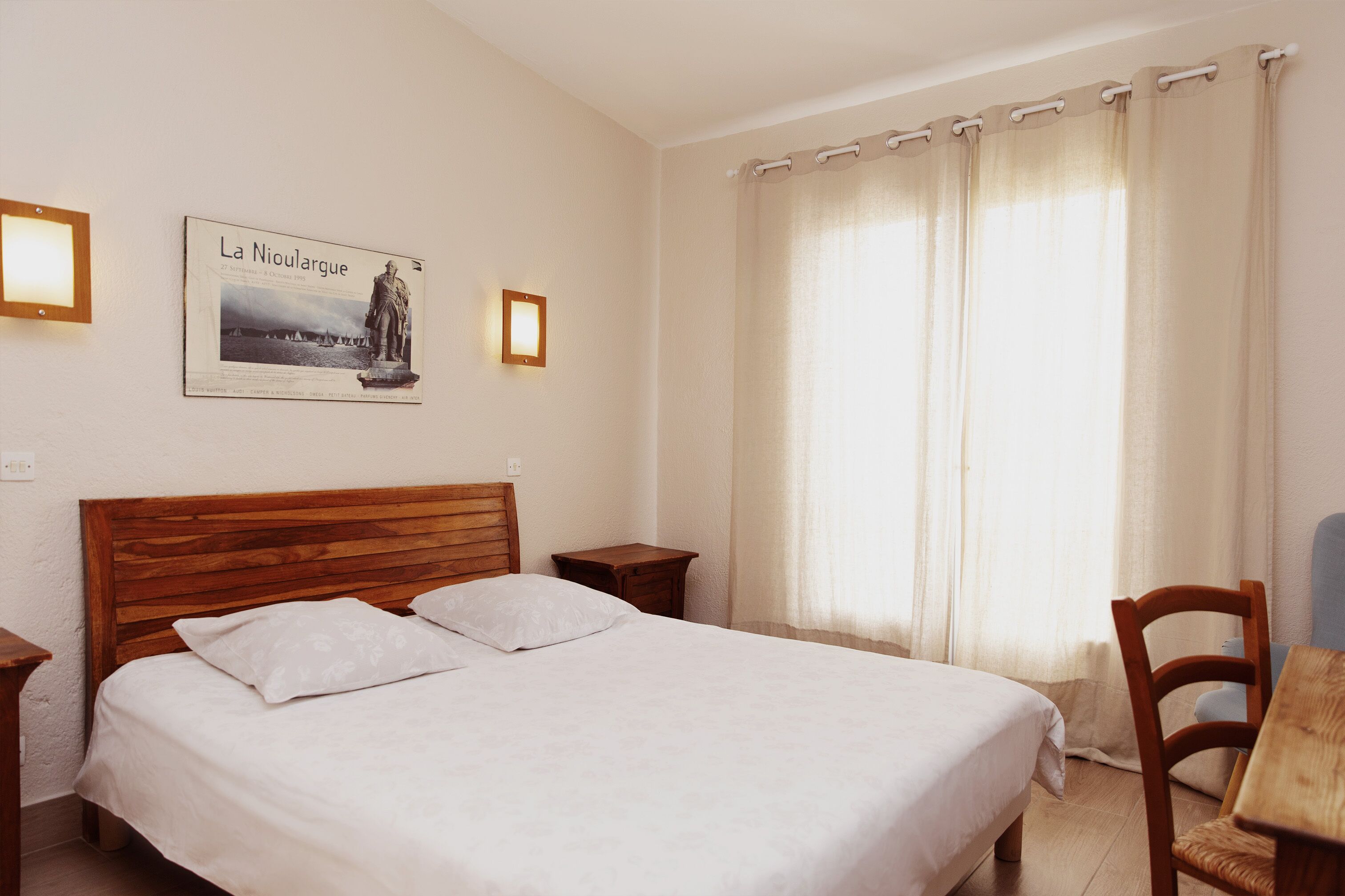 A neatly made bed in a room with a wooden headboard, a small wooden nightstand, a framed picture on the wall, and a window with curtains.