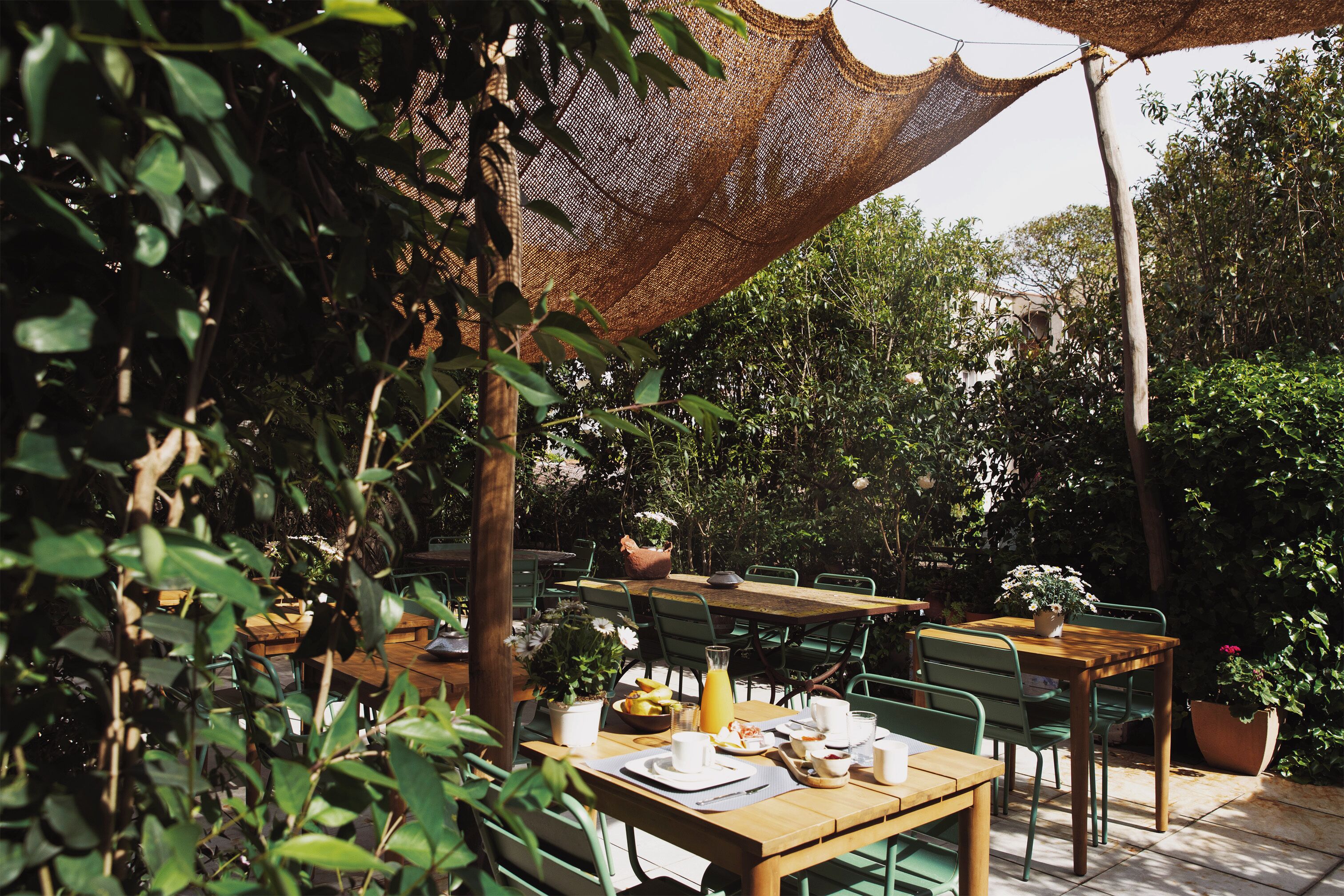 An outdoor dining area with wooden tables and chairs, surrounded by greenery and potted plants.