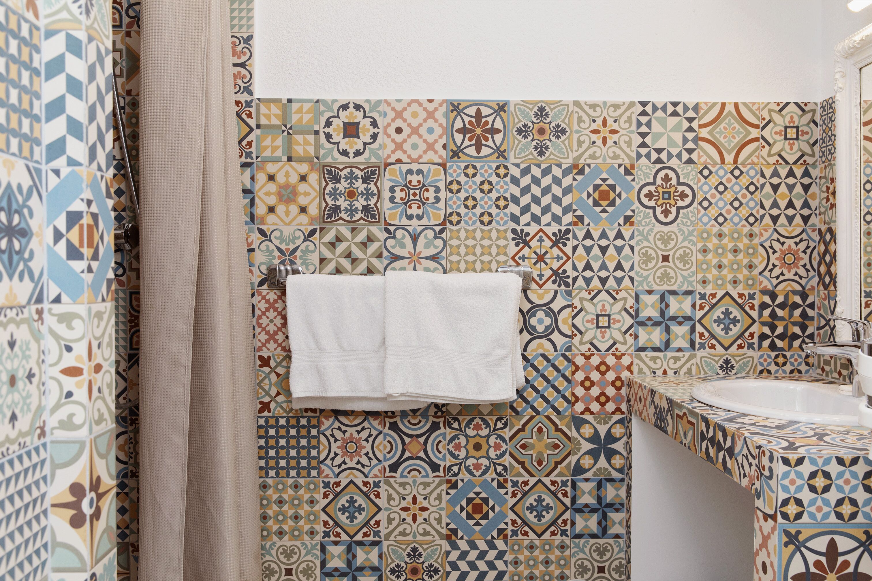 Un baño con un muro de azulejos que presenta una variedad de patrones geométricos y florales. Hay una toalla blanca colgada en la pared, un lavamanos y una cortina de ducha.
