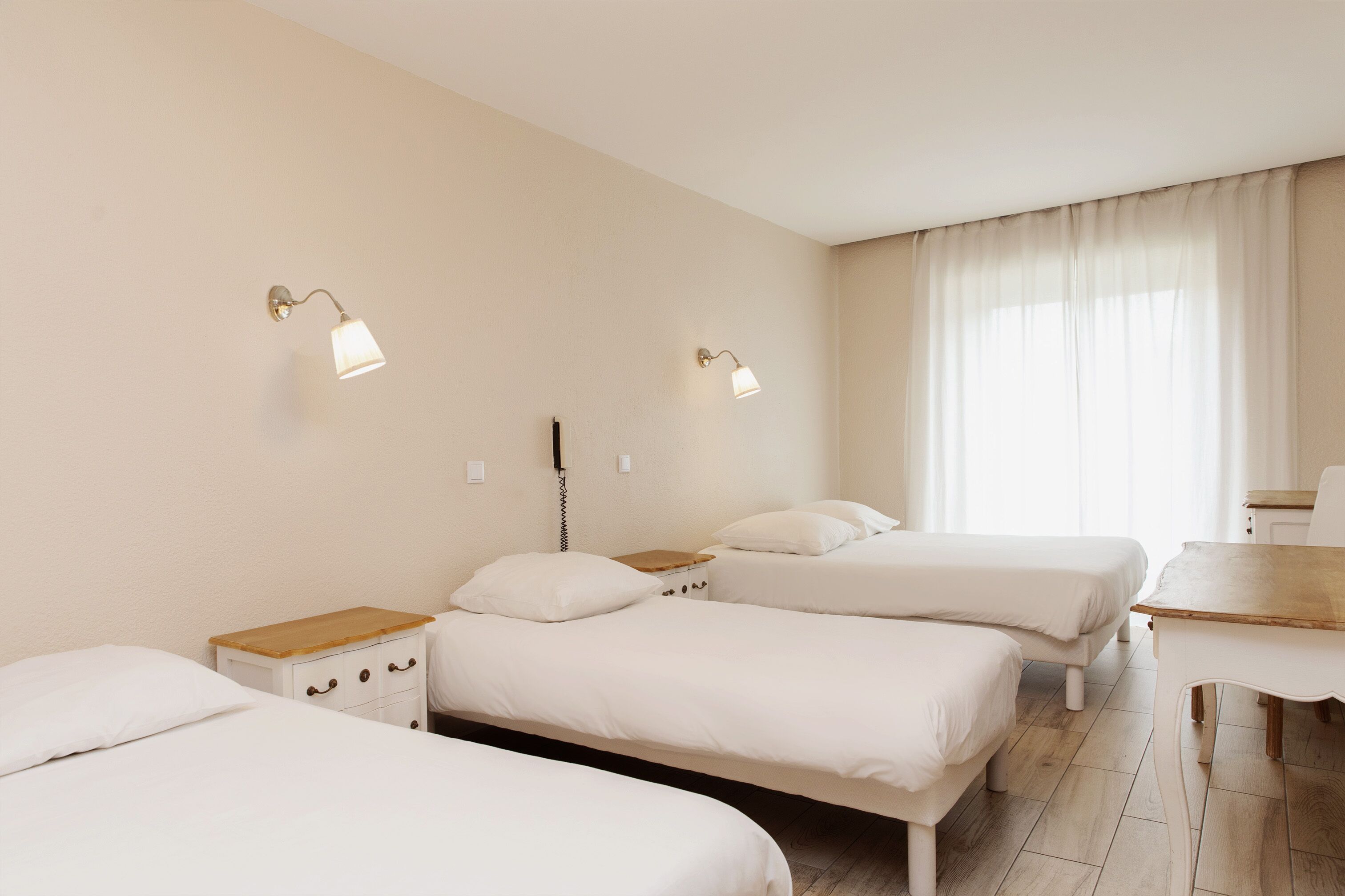 A hotel room with three single beds, a wooden nightstand, a desk, and two wall-mounted lamps.