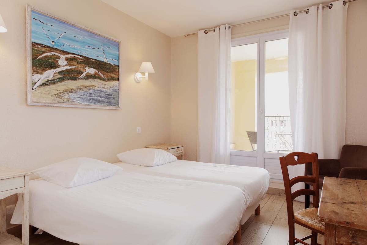 A hotel room with two beds, a wooden desk, a chair, a table, a painting of birds, and a balcony with curtains.