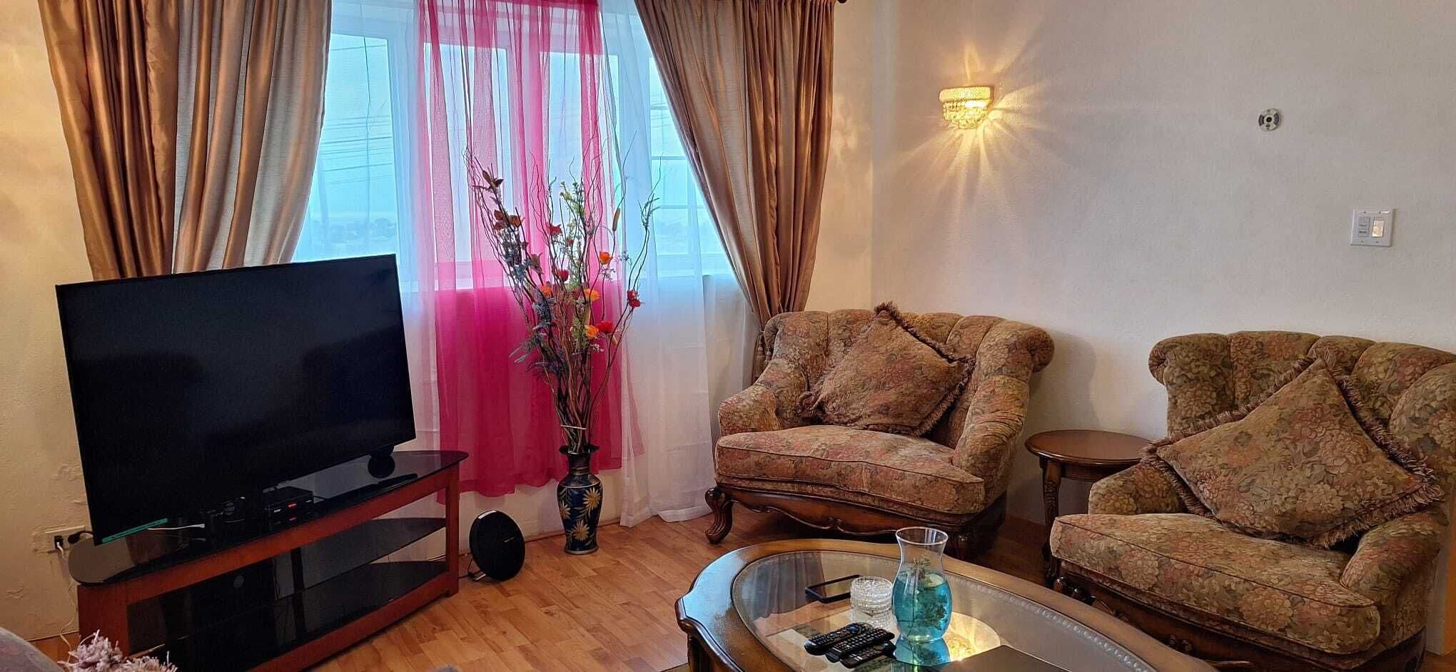 Ein Wohnzimmer mit einem Flachbildfernseher auf einem Holzboden, zwei gemusterten Sesseln, einem kleinen Couchtisch und einer Vase mit Blumen.