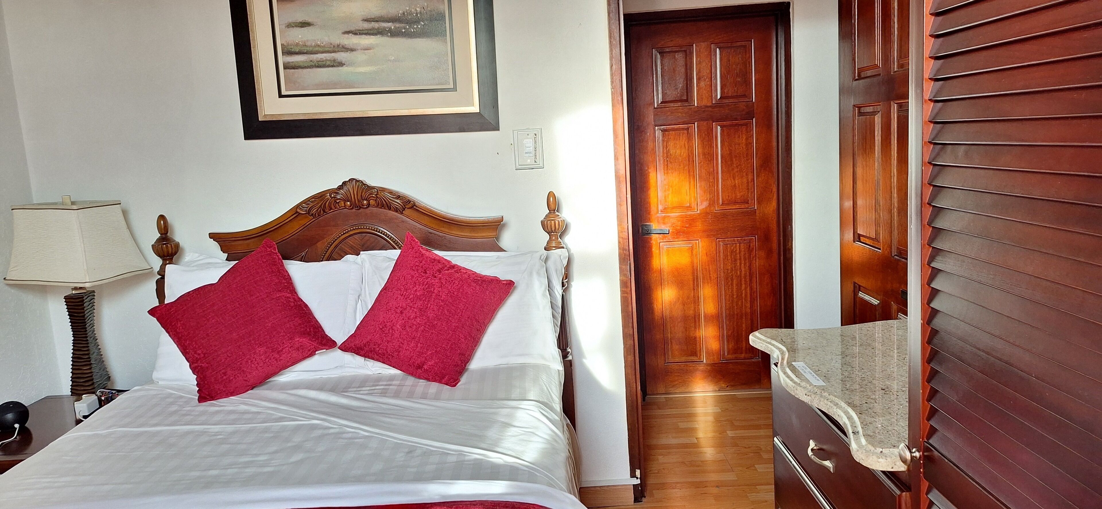 A neatly made bed with white and red pillows, a wooden headboard, a bedside lamp, and a framed picture on the wall.