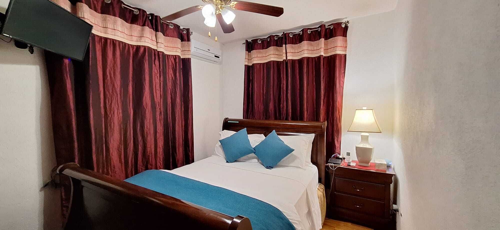 Un dormitorio con una cama de madera, dos almohadas azules, una lámpara de noche y un televisor montado en la pared.