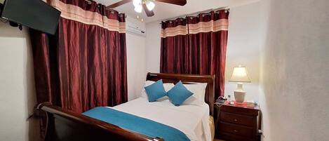 Un dormitorio con una cama de madera, dos almohadas azules, una lámpara de noche y un televisor montado en la pared.