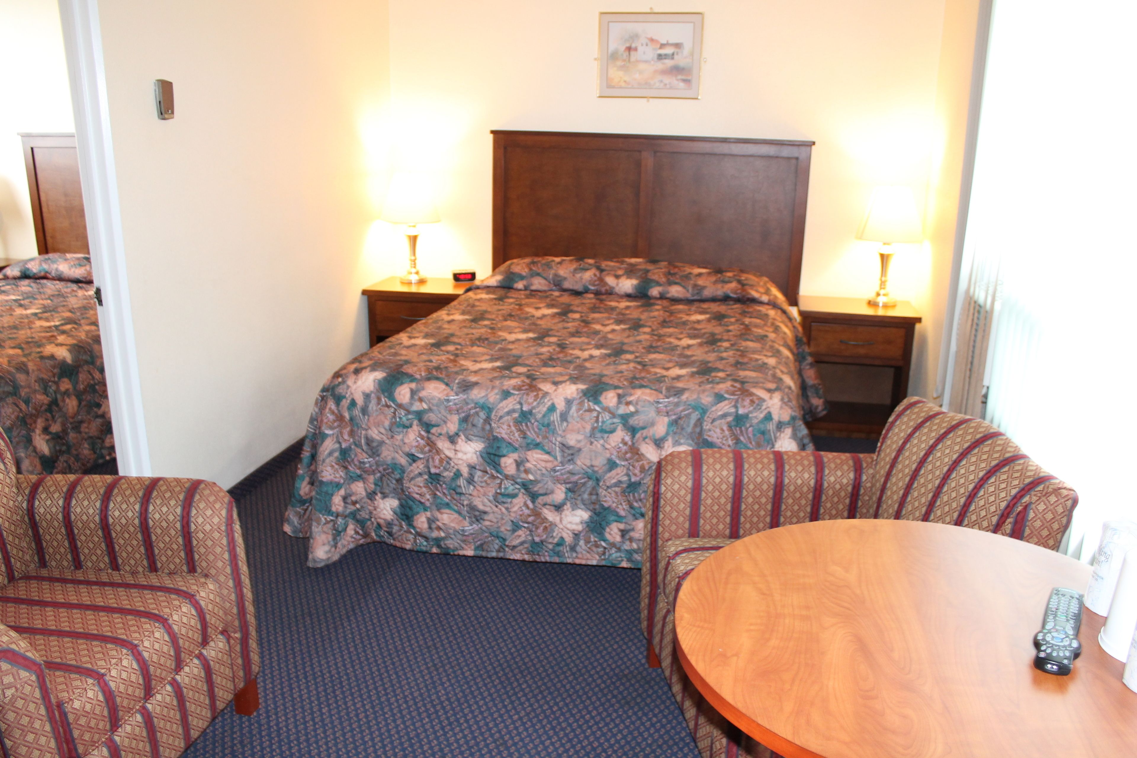A hotel room with a large bed, two bedside tables with lamps, a round wooden table, and two patterned armchairs.
