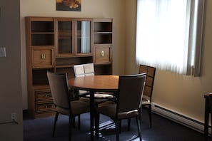 Un comedor con una mesa redonda y cuatro sillas, armarios de madera con puertas de cristal, y una ventana con persianas.