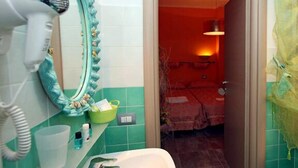 Ein Badezimmer mit Fön, Spiegel und Waschbecken. Eine Tür führt zu einem Raum mit Bett und Lampe.