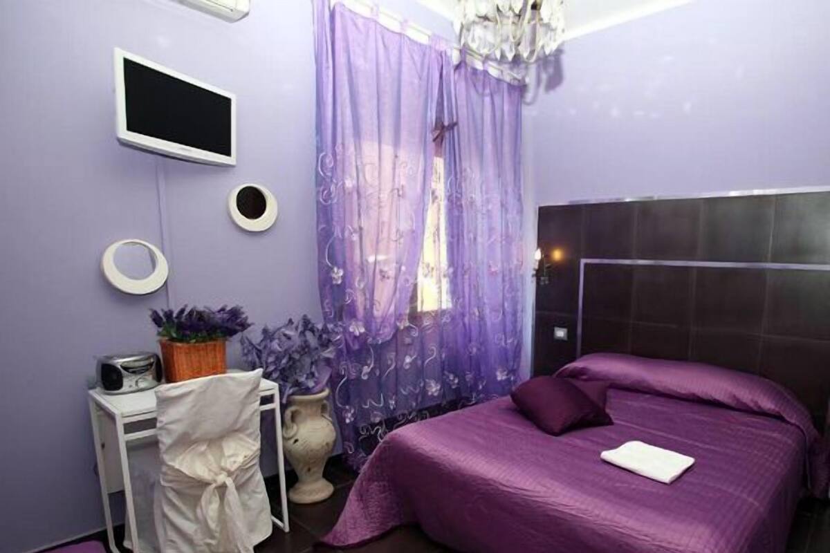 A bedroom with a bed, a desk, a chair, a TV, and a window with curtains.