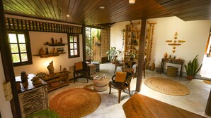 A cozy living room with wooden furniture, a large woven rug, and a bookshelf filled with decorative items. - Beco Pousada Boutique Noronha (Fernando de Noronha)