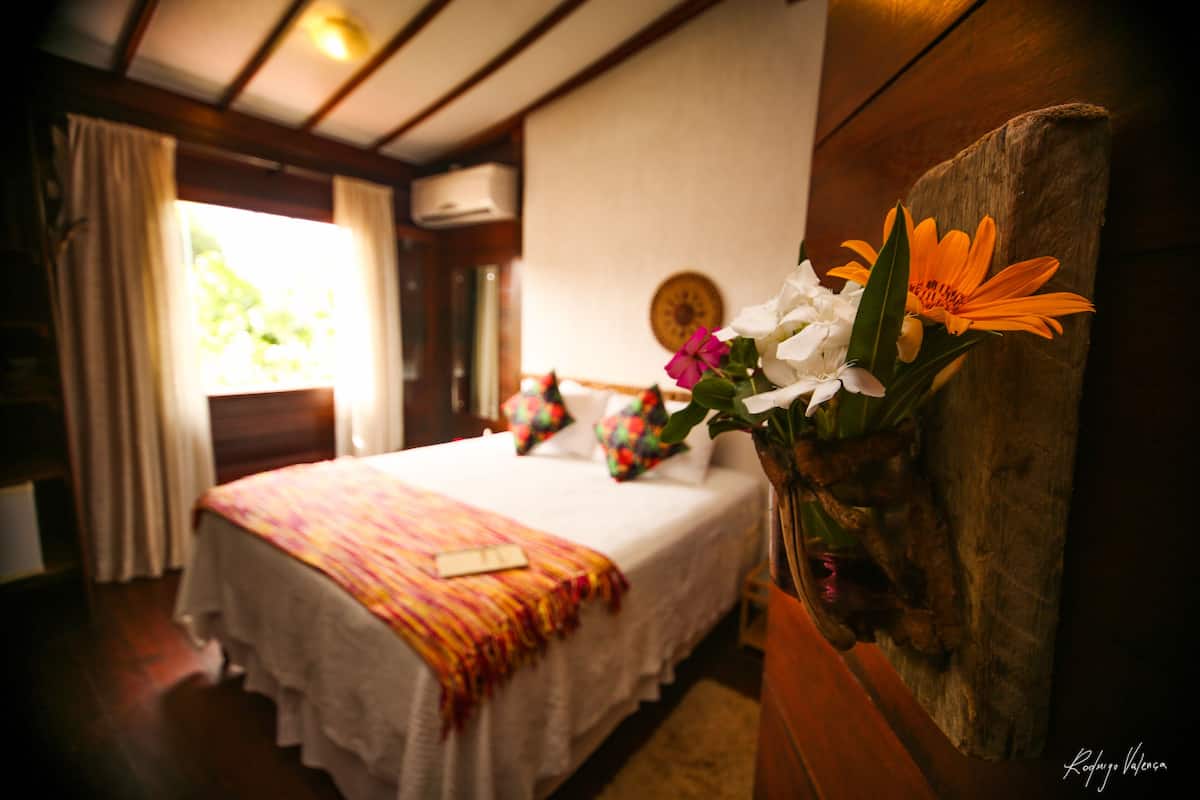 A hotel room with a bed, a window with curtains, a wall clock, and a vase with flowers.
