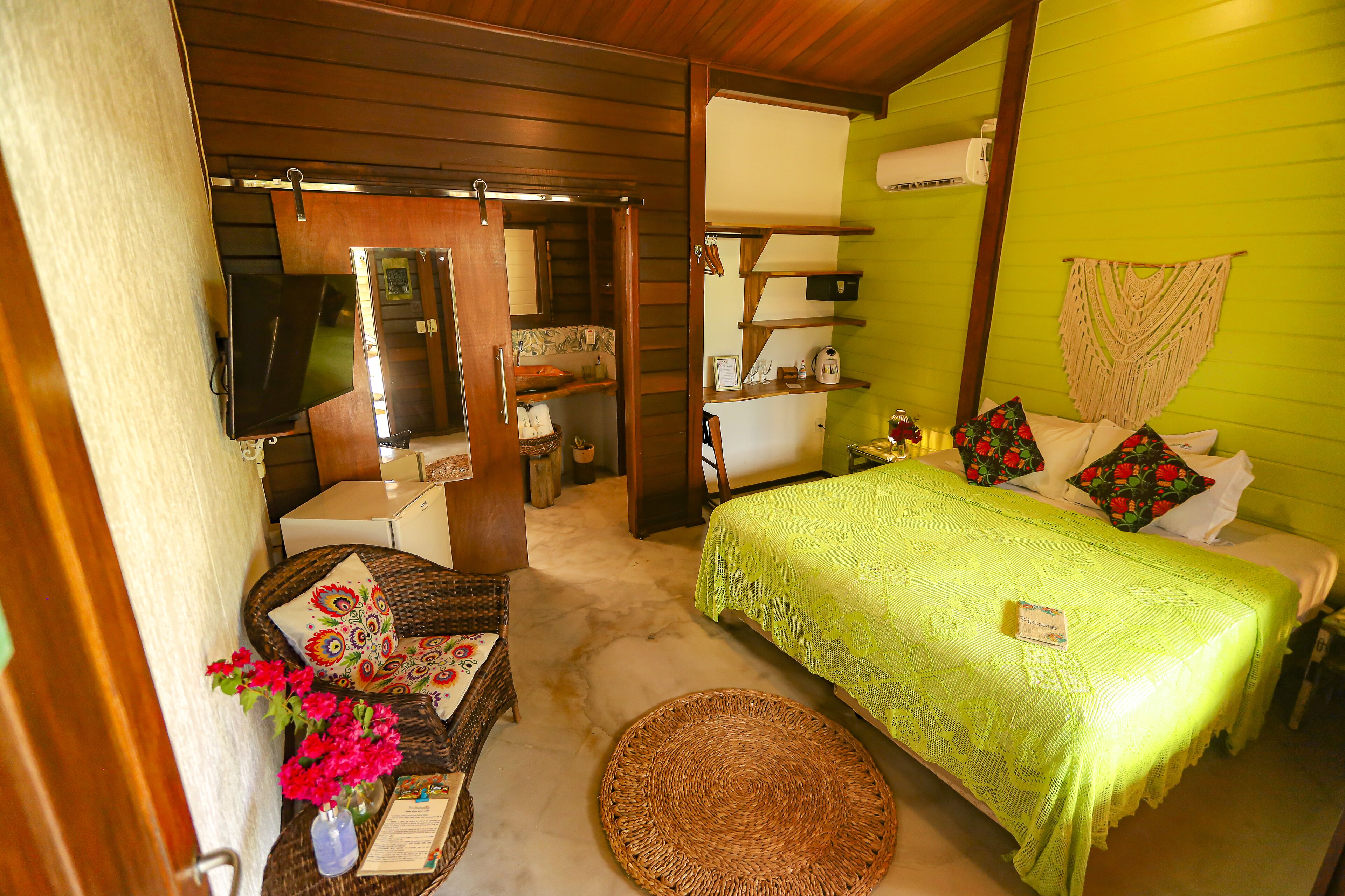 Una habitación con una cama, una silla, una mesita con flores, un televisor y un espejo.