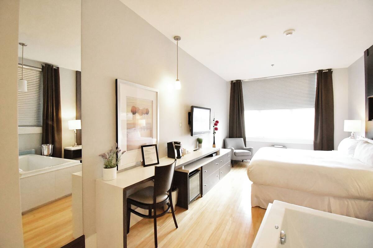 A modern hotel room with a bed, a desk with a chair, a television, and a window with curtains.