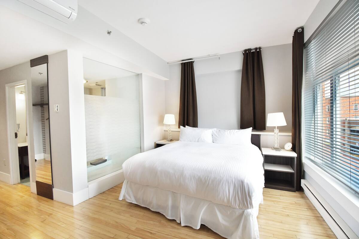 A modern hotel room with a large bed, bedside tables, a glass shower enclosure, and a view of the city through the window.
