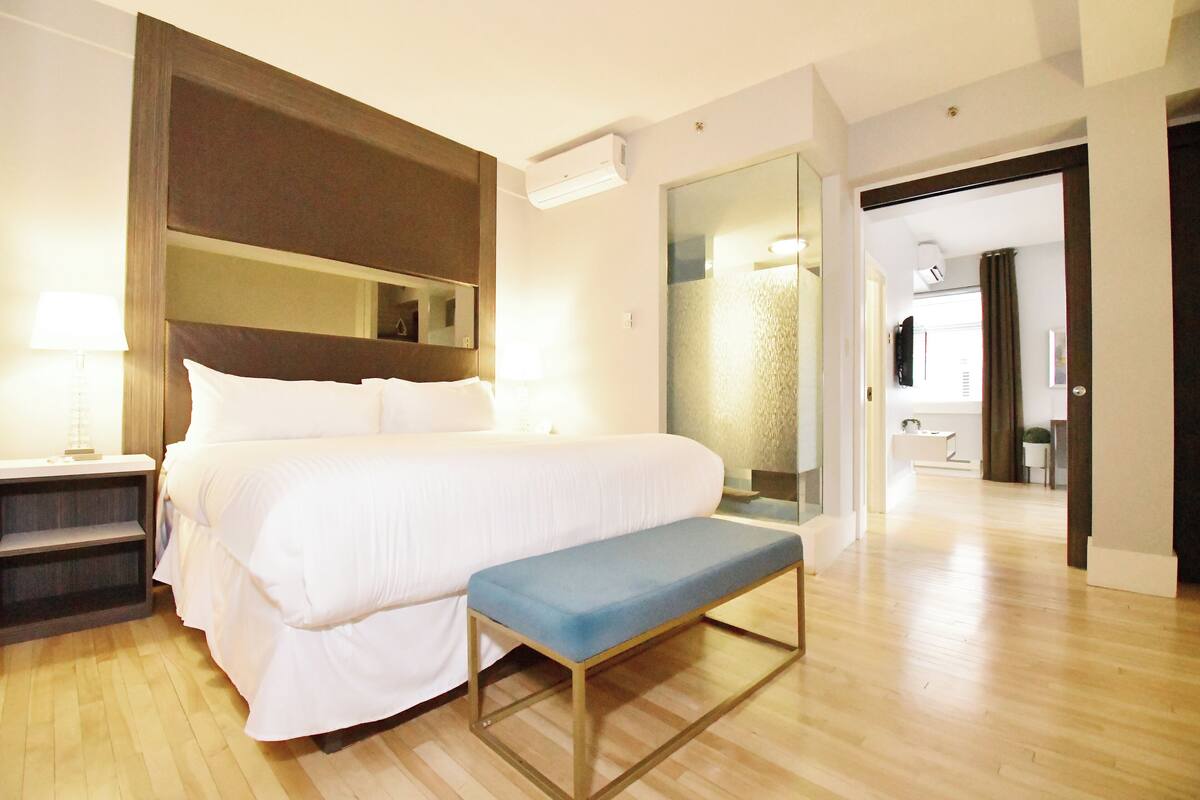 A hotel room with a large bed, a bedside table, a bench, a mirror, and a bathroom visible through an open door.
