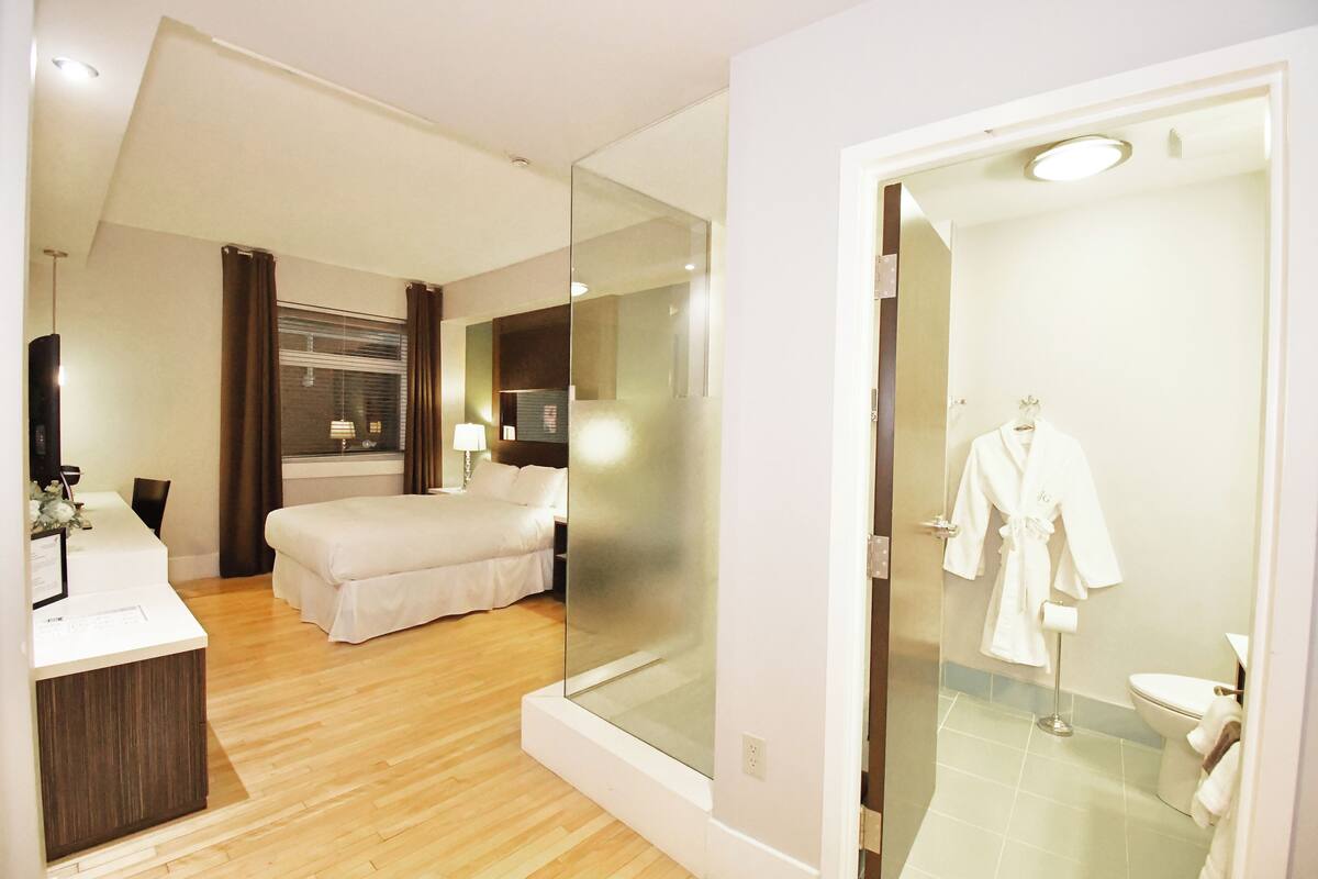 A hotel room with a wooden floor, a bed, a desk with a mirror, a bathroom with a glass shower enclosure, and a robe hanging on the wall.