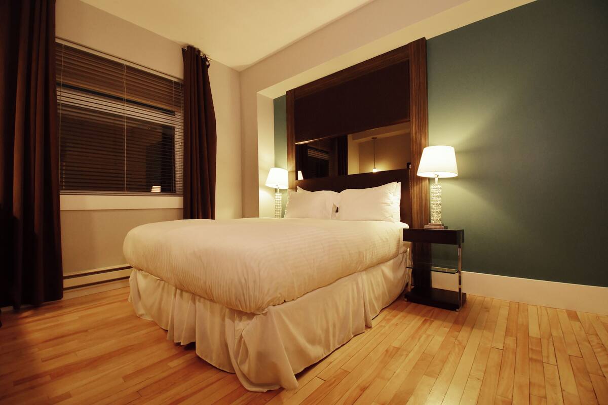 A neatly made bed in a hotel room with a wooden floor, a nightstand with lamps, and a large window with curtains.