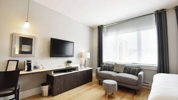 A modern hotel room with a flat-screen TV, a sofa, a small ottoman, a desk with a coffee maker, and a window with blinds.