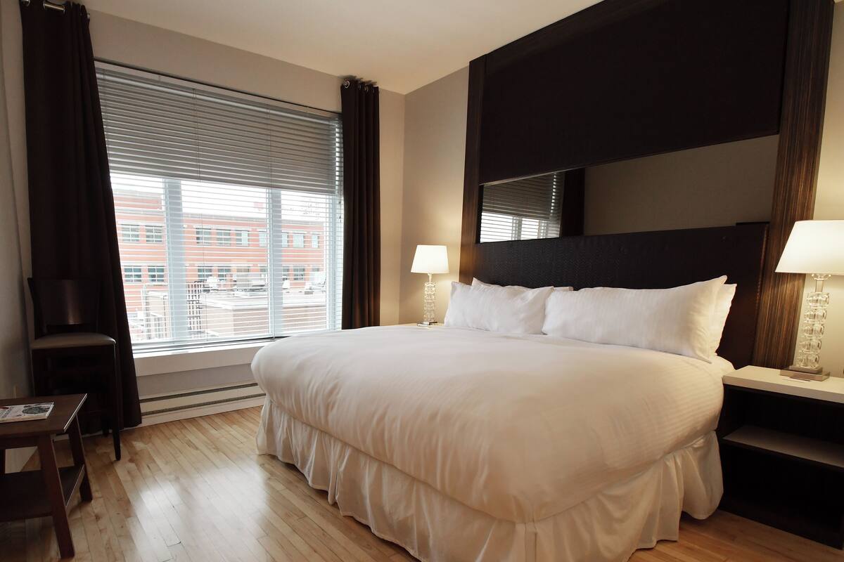 A large bed with white linens, a wooden floor, a nightstand with a lamp, and a window with blinds.