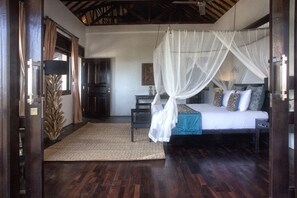 A bedroom with a canopy bed, a wooden floor, and a door with a curtain. - Villa Flow (Karangasem)