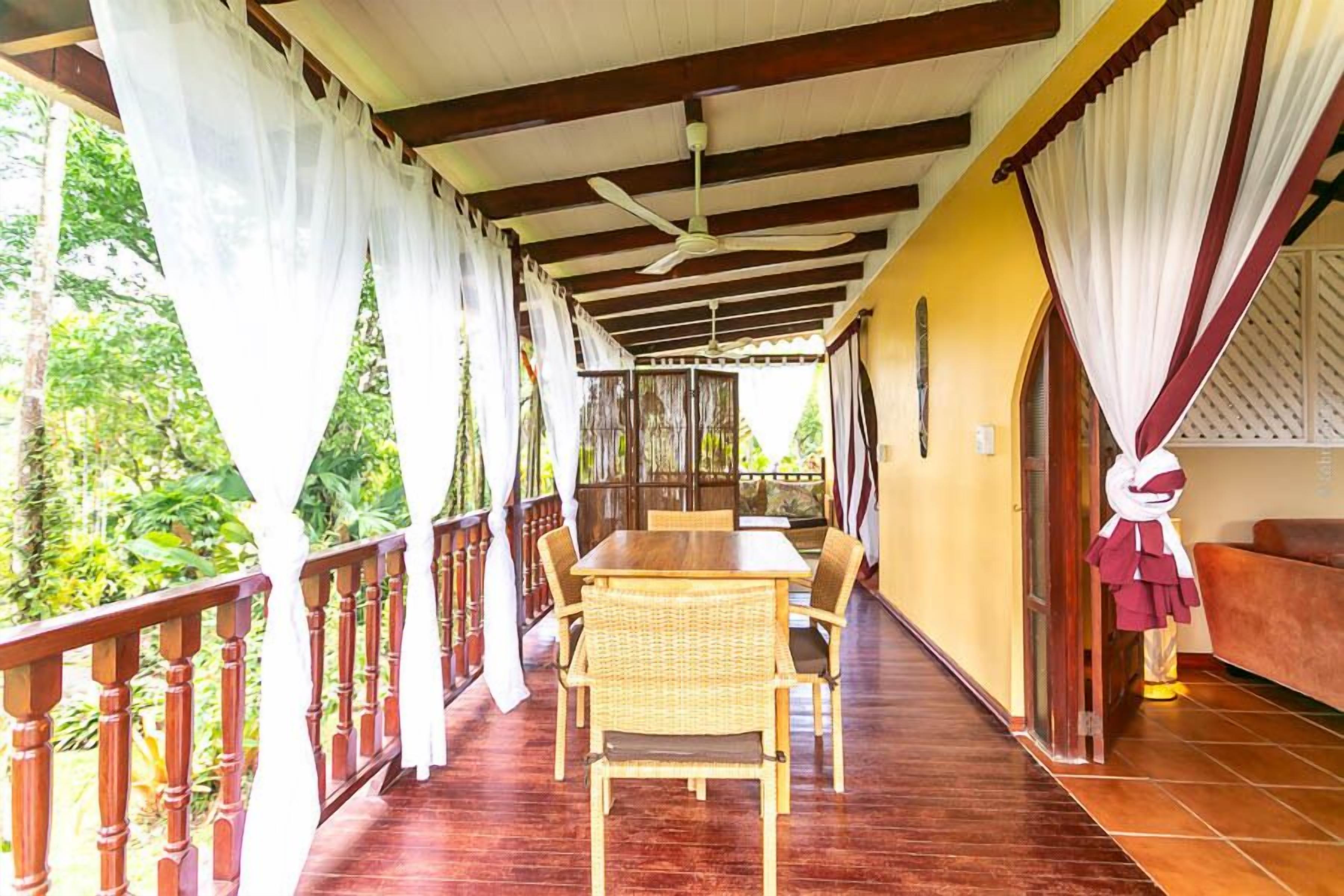 A covered outdoor area with a wooden table and chairs, a ceiling fan, and a view of greenery.