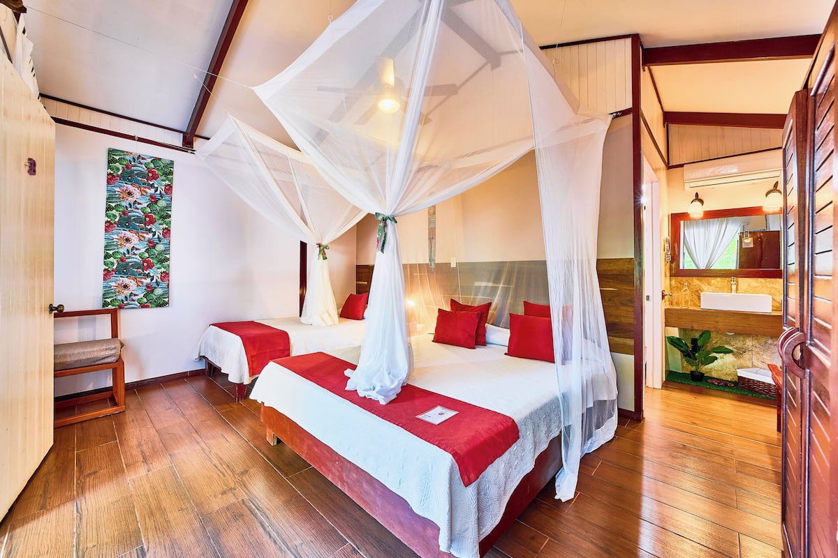 A bedroom with two beds, a wooden floor, a canopy bed, a painting, and a bathroom with a sink and mirror.