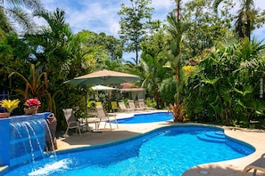 Piscina com cascata, cercada por vegetação exuberante e cadeiras de descanso.